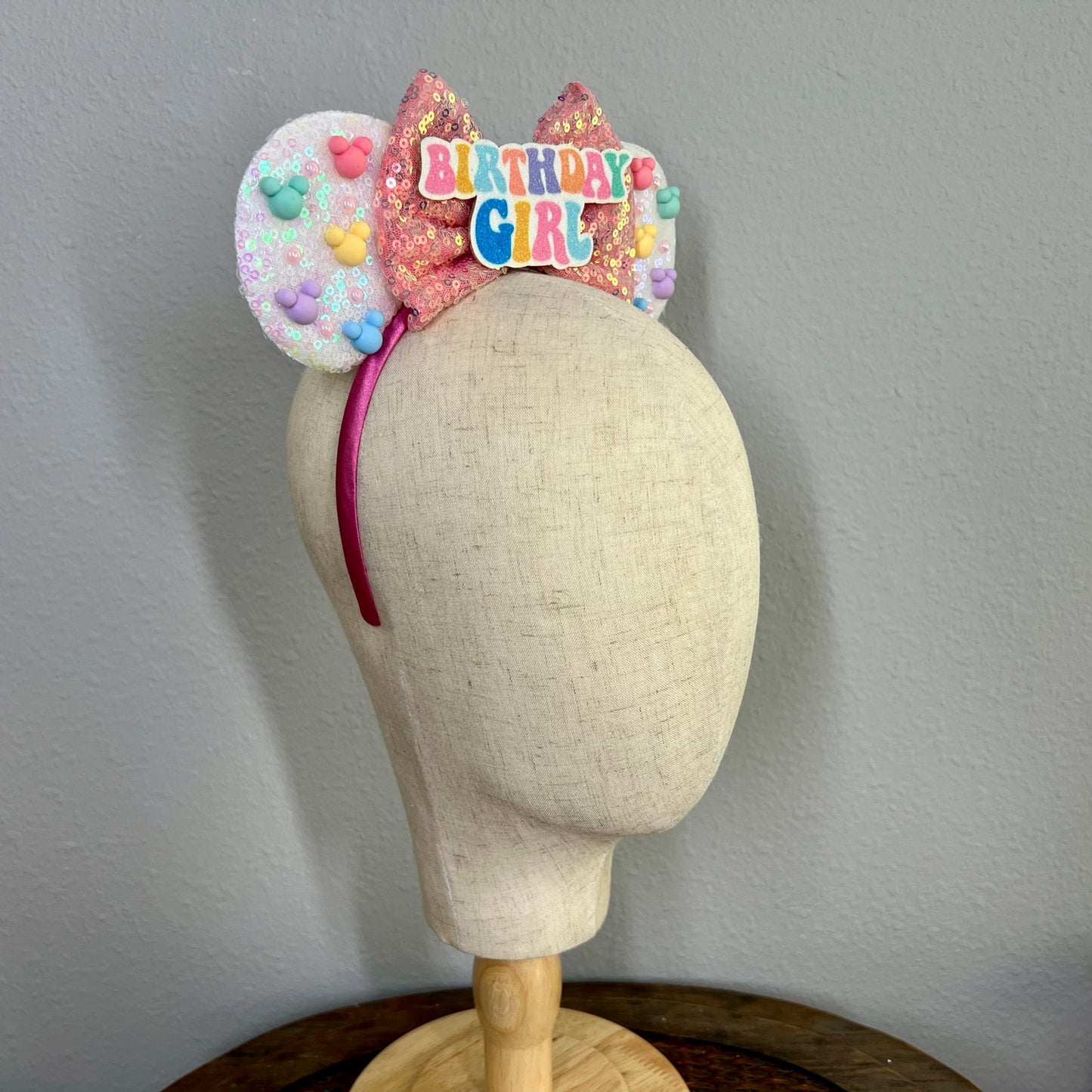 Mannequin head wearing white sequin headband with pink bow, rainbow mouse-shaped pearls, and glitter "Birthday Girl" text centerpiece