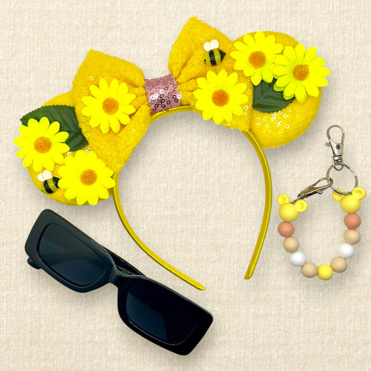 Yellow floral mouse ears headband with yellow daisies and bees and a bow; black sunglasses; yellow beaded mouse ears holder on a beige background