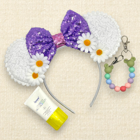 Mouse ears headband with purple sequin bow and white flowers; small bottle of sunscreen; colorful beaded mouse ears holder on a beige background
