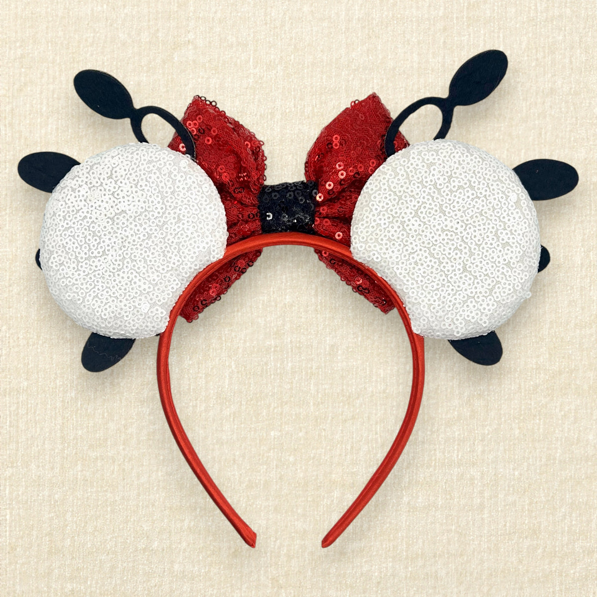 A Minnie Mouse-inspired sequin headband featuring two white sequined circular ears with black accents, a red sequin bow, and a red headband, displayed on a textured beige background.