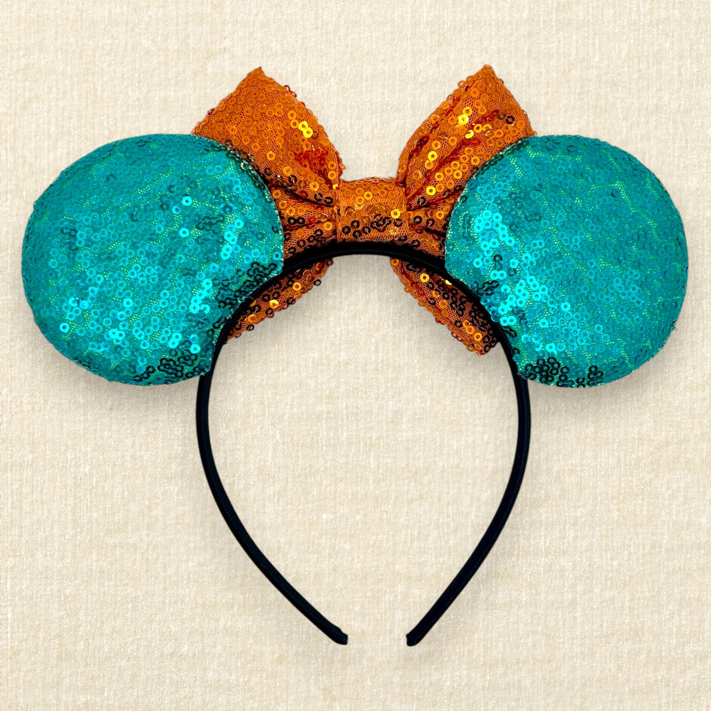 Halloween-themed mouse ears headband with orange sequin bow, black bats, and teal blue ears on a beige background (back view)