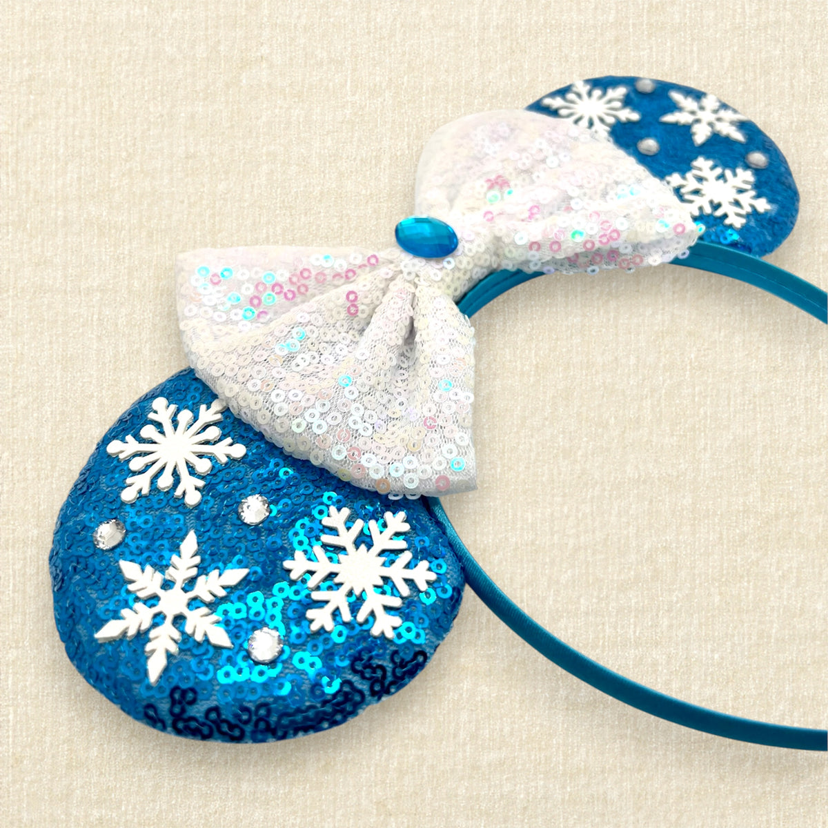 Winter-themed blue sequin mouse ears headband with white snowflakes, an iridescent white sequin bow, and blue and crystal rhinestone accents.