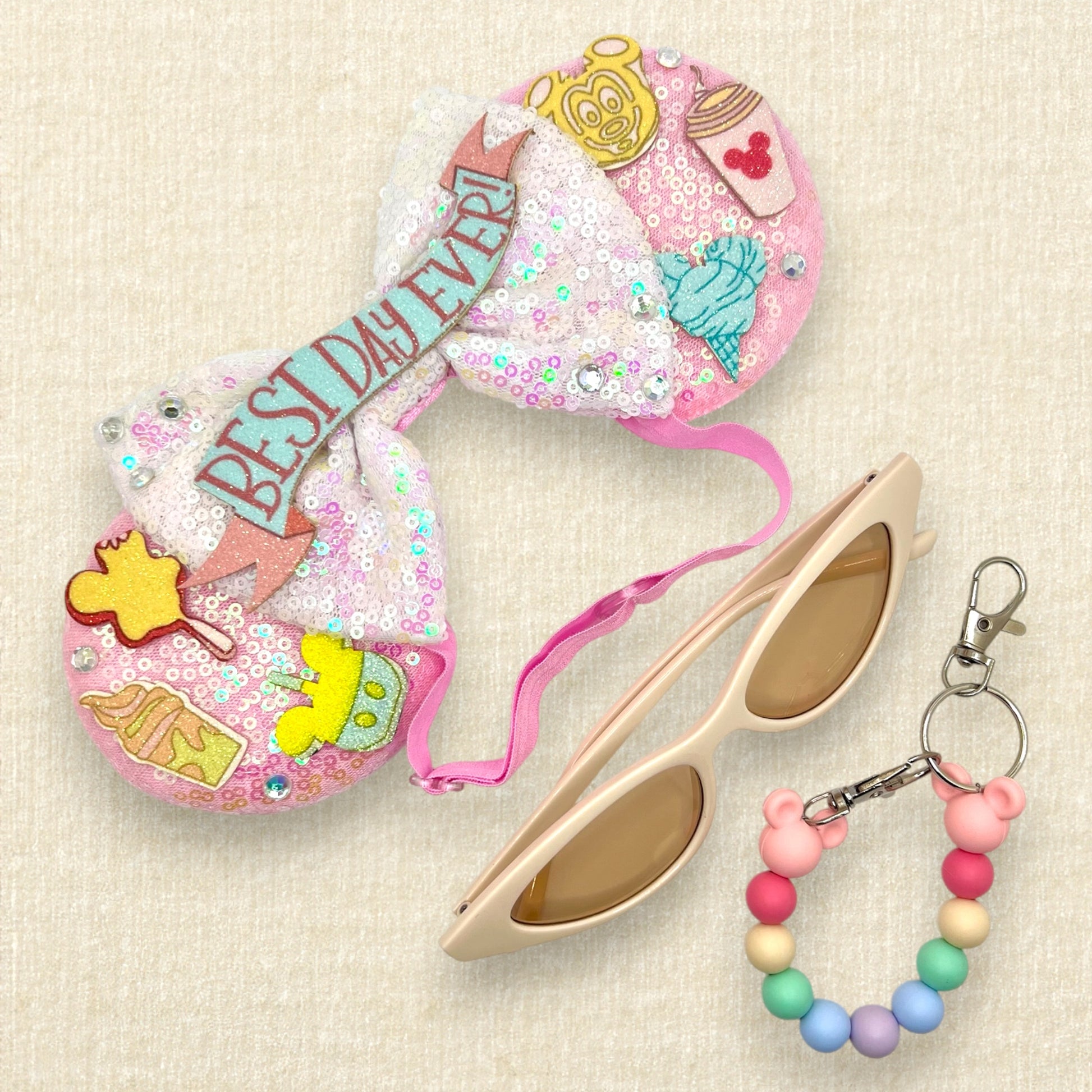 Pink mouse ears with glitter theme park treats, iridescent white bow, and glitter "Best Day Ever!" text on an elastic headband; cream cat eye sunglasses; colorful beaded mouse ears holder keychain