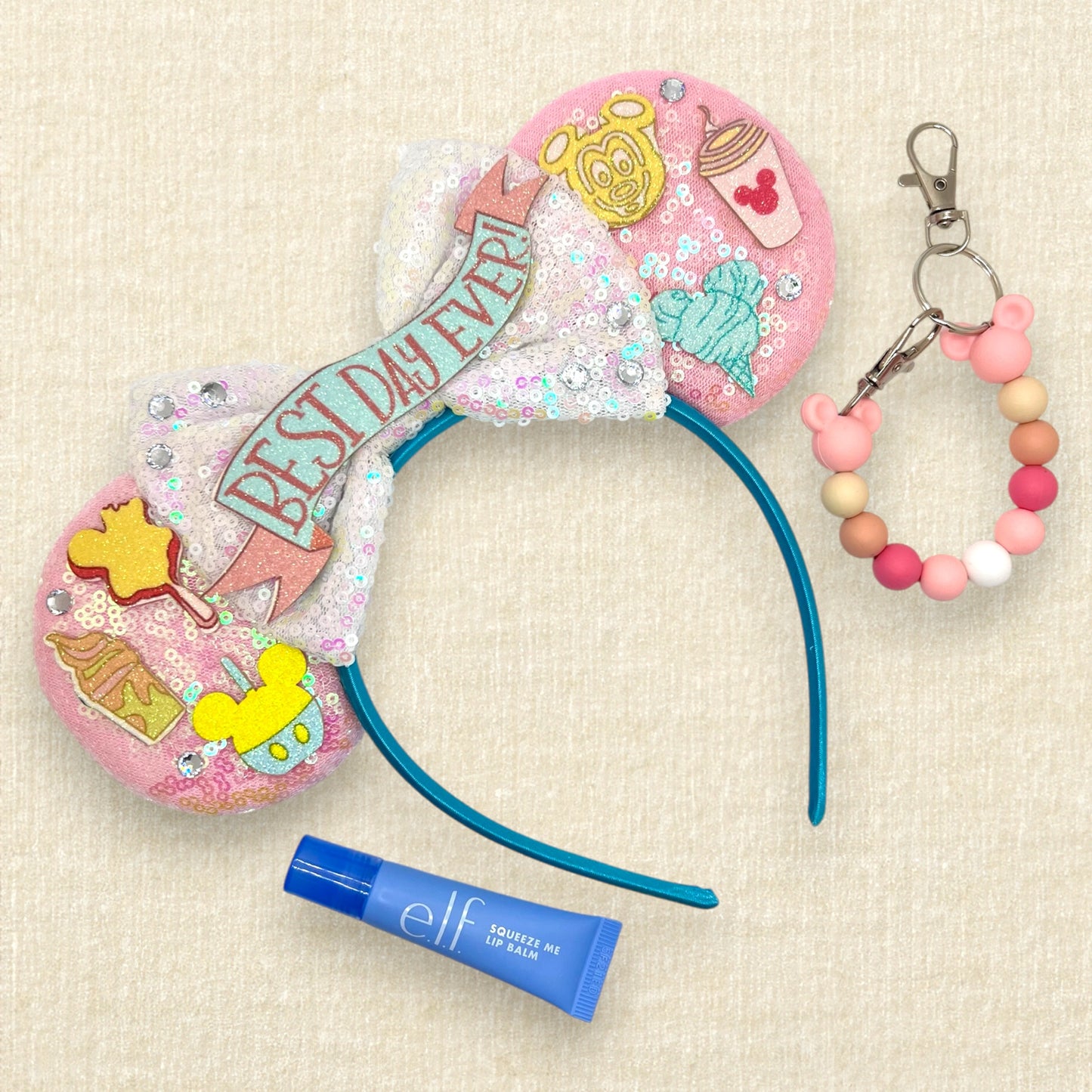 Pink mouse ears headband with glitter theme park snack details, iridescent white bow, and glitter "Best Day Ever!" text; lip balm; pink beaded mouse ears holder keychain