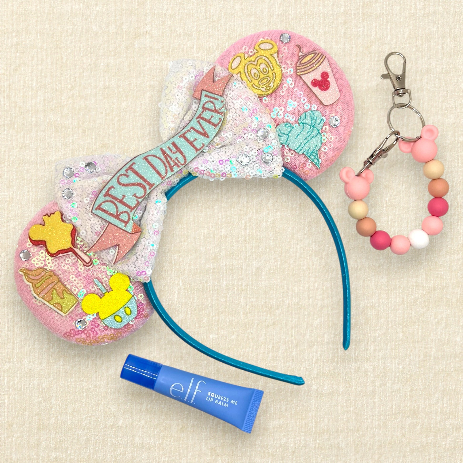 Pink mouse ears headband with glitter theme park snack details, iridescent white bow, and glitter "Best Day Ever!" text; lip balm; pink beaded mouse ears holder keychain