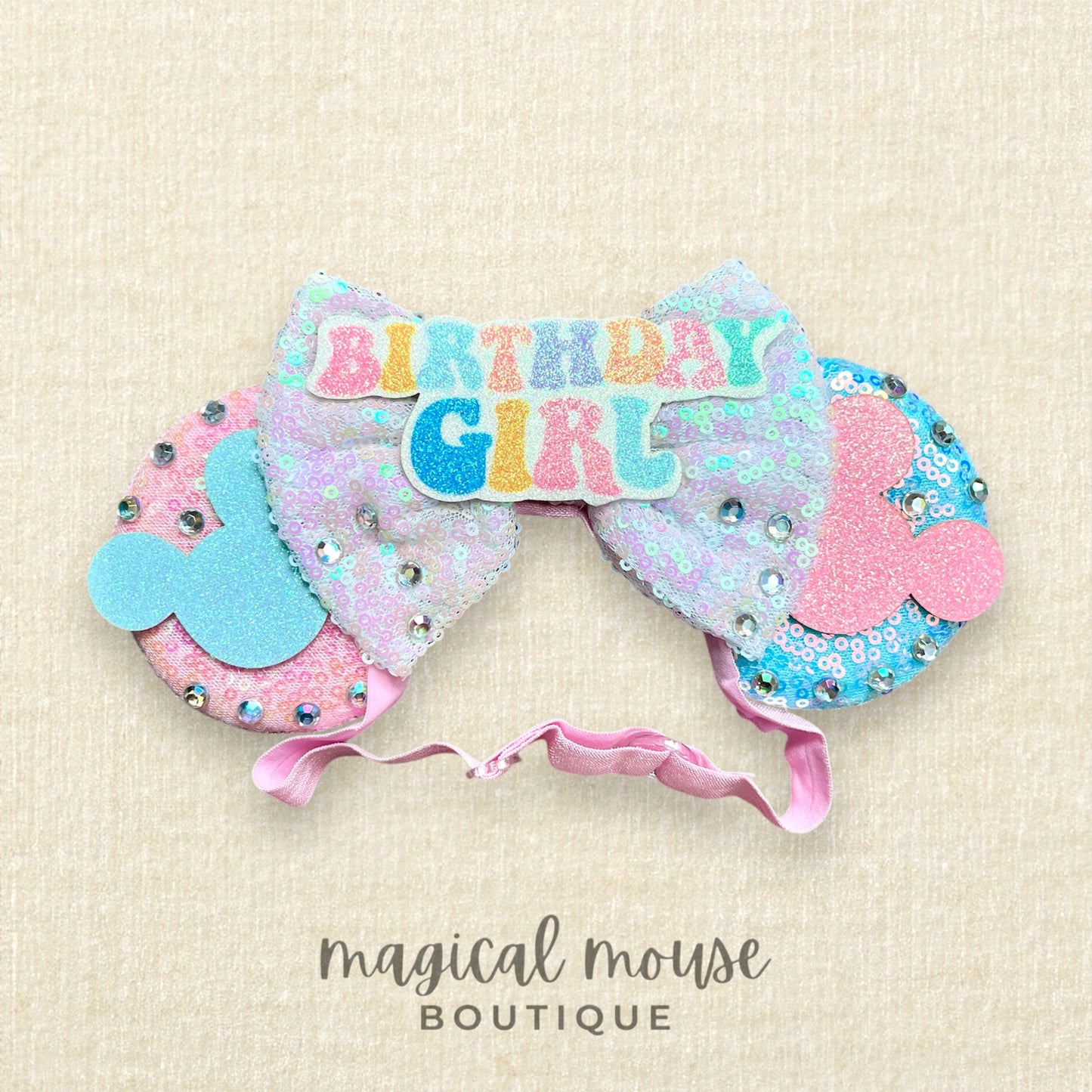 Pink and blue elastic mouse ears headband with 'Birthday Girl' text centerpiece on a beige background with 'magical mouse boutique' branding