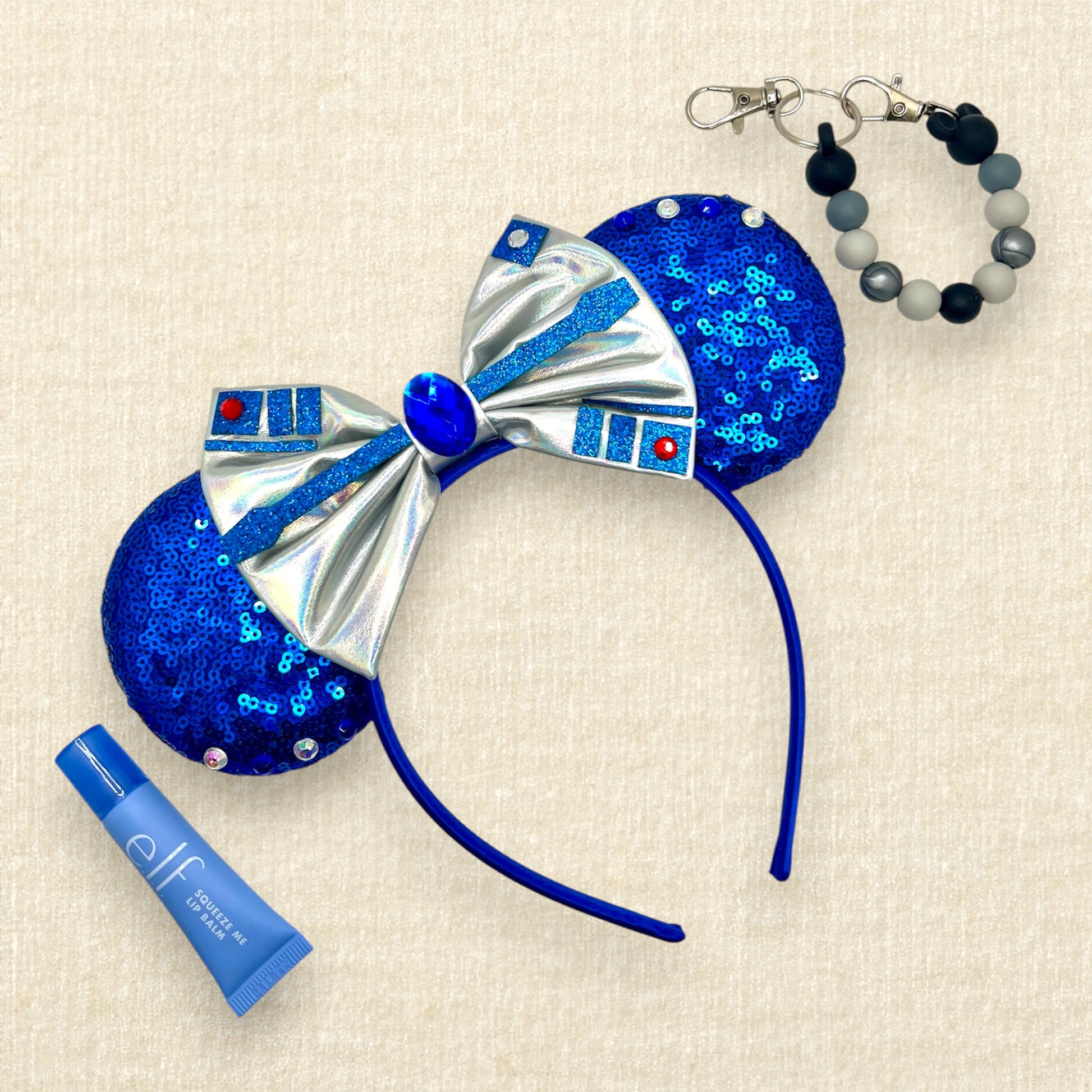 Blue mouse ears with metallic silver bow, rhinestones, and glitter robot-inspired details with blue lip gloss and monochrome mouse ears holder