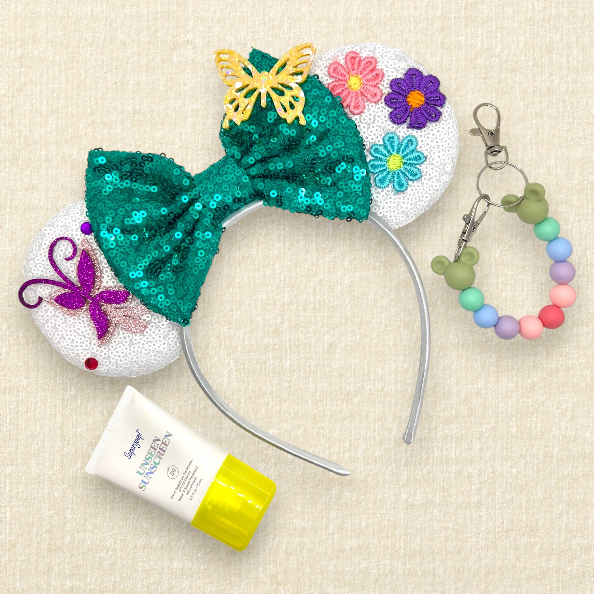 White sequin mouse ears with butterflies and flower details and a green sequin bow; sunscreen; colorful beaded mouse ears holder keychain
