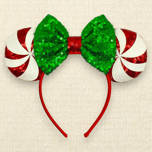 Holiday-themed candy cane ears headband featuring green sequined bows and red-and-white striped candy cane ears on a red headband, displayed on a textured beige background.