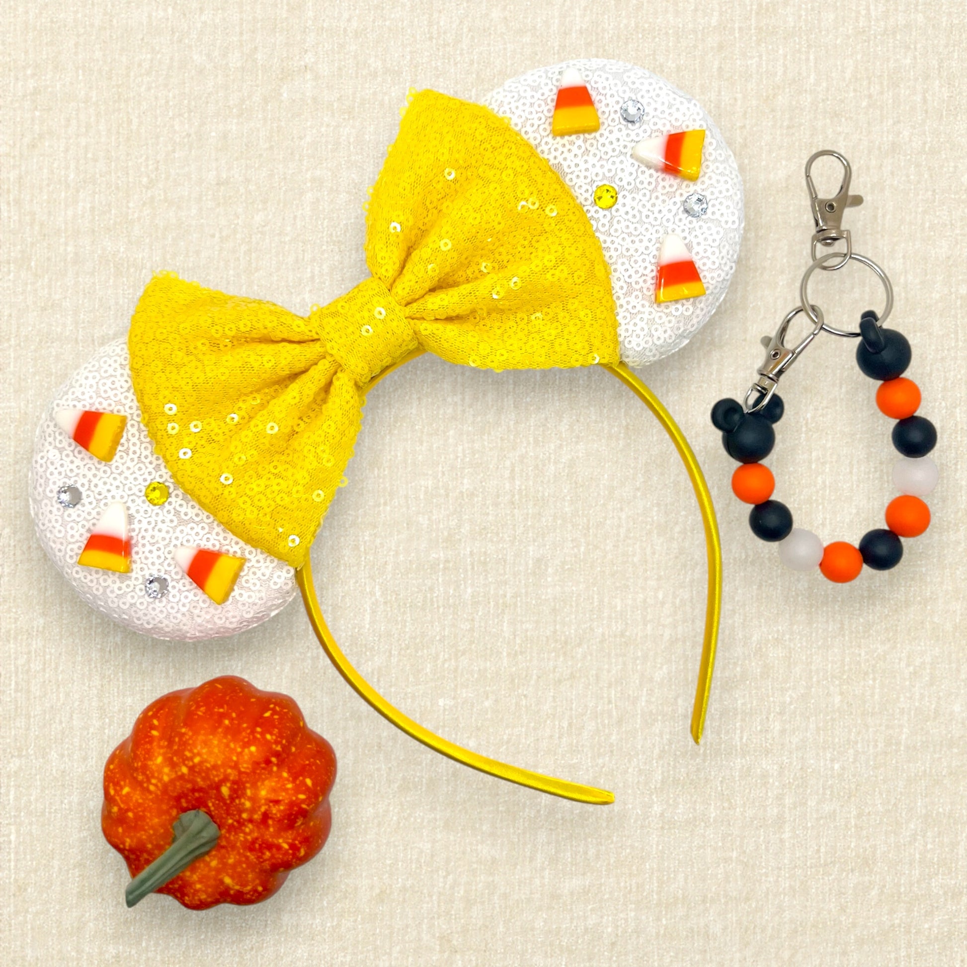Halloween-themed mouse ears headband with white ears, candy corn pieces, rhinestones, and a yellow bow; orange pumpkin decoration; black and orange beaded mouse ears holder keychain