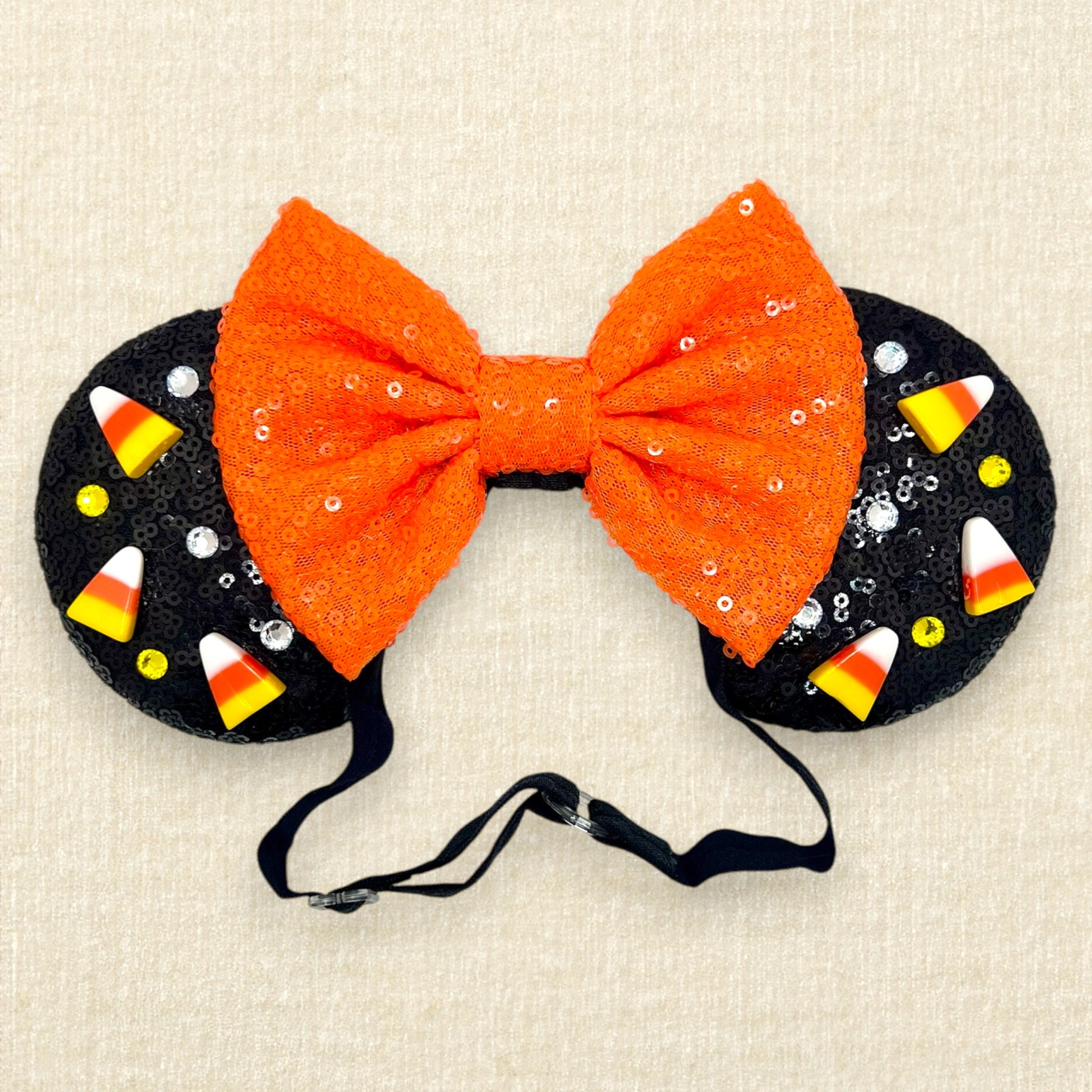 Black sequin Halloween-themed mouse ears with candy corn pieces, rhinestones, and an orange bow on an elastic headband