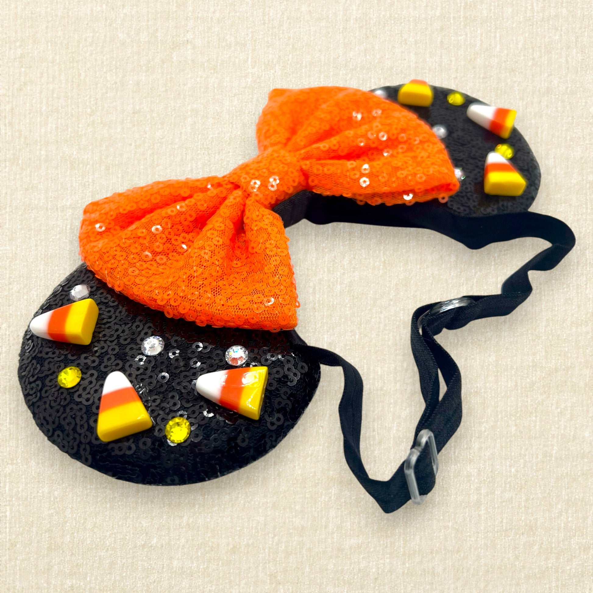 Black sequin Halloween-themed mouse ears with candy corn pieces, rhinestones, and an orange bow on an elastic headband