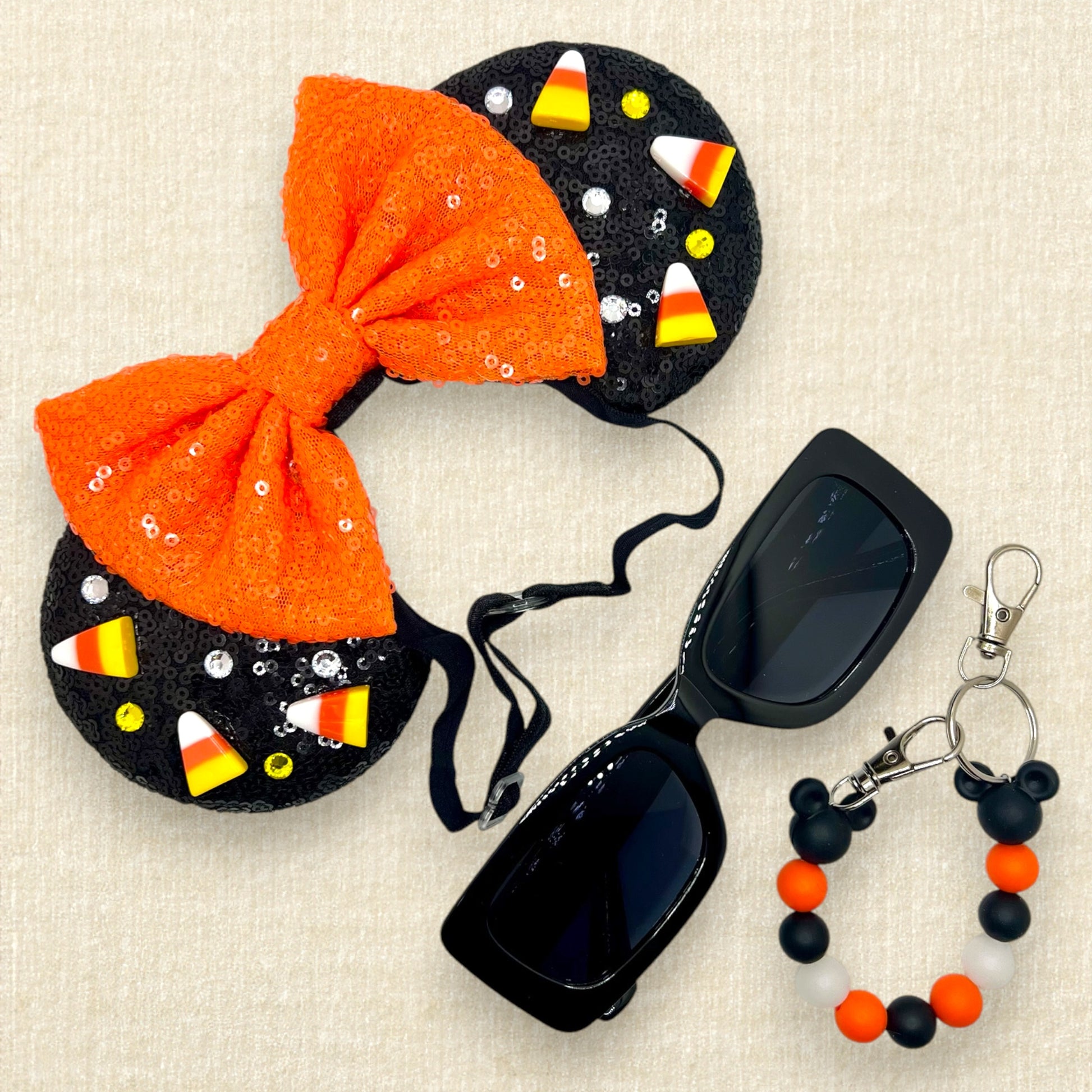 Black sequin Halloween-themed mouse ears with candy corn pieces, rhinestones, and an orange bow on an elastic headband; black sunglasses; orange and black beaded mouse ears holder keychain