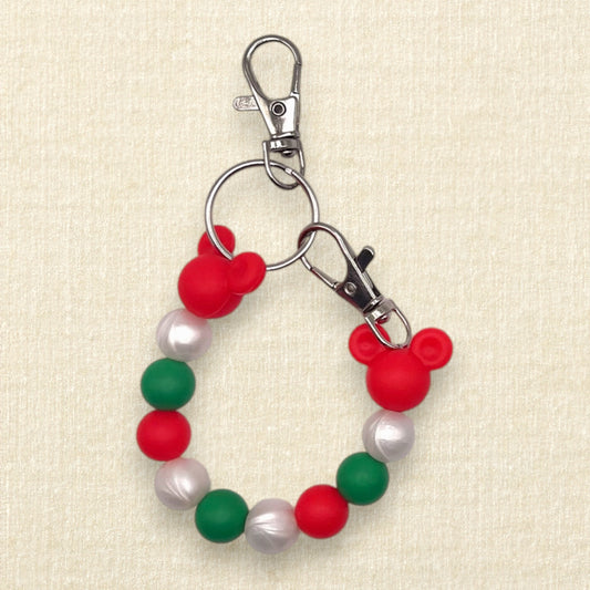 A Christmas-themed beaded mouse ears holder keychain featuring red and green silicone beads, white faux-pearl beads, and red Mickey Mouse head silhouettes, attached to silver metal clasps on a textured beige background.