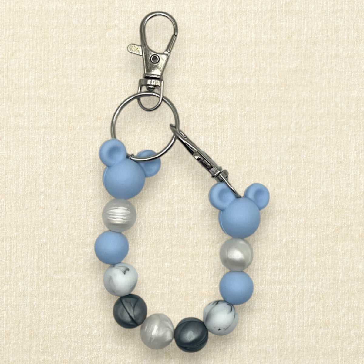 A winter-themed beaded mouse ears holder keychain featuring light blue silicone Mickey Mouse ears, light blue and gray beads, and marbled black and white beads, attached to a silver metal clasp on a textured beige background.