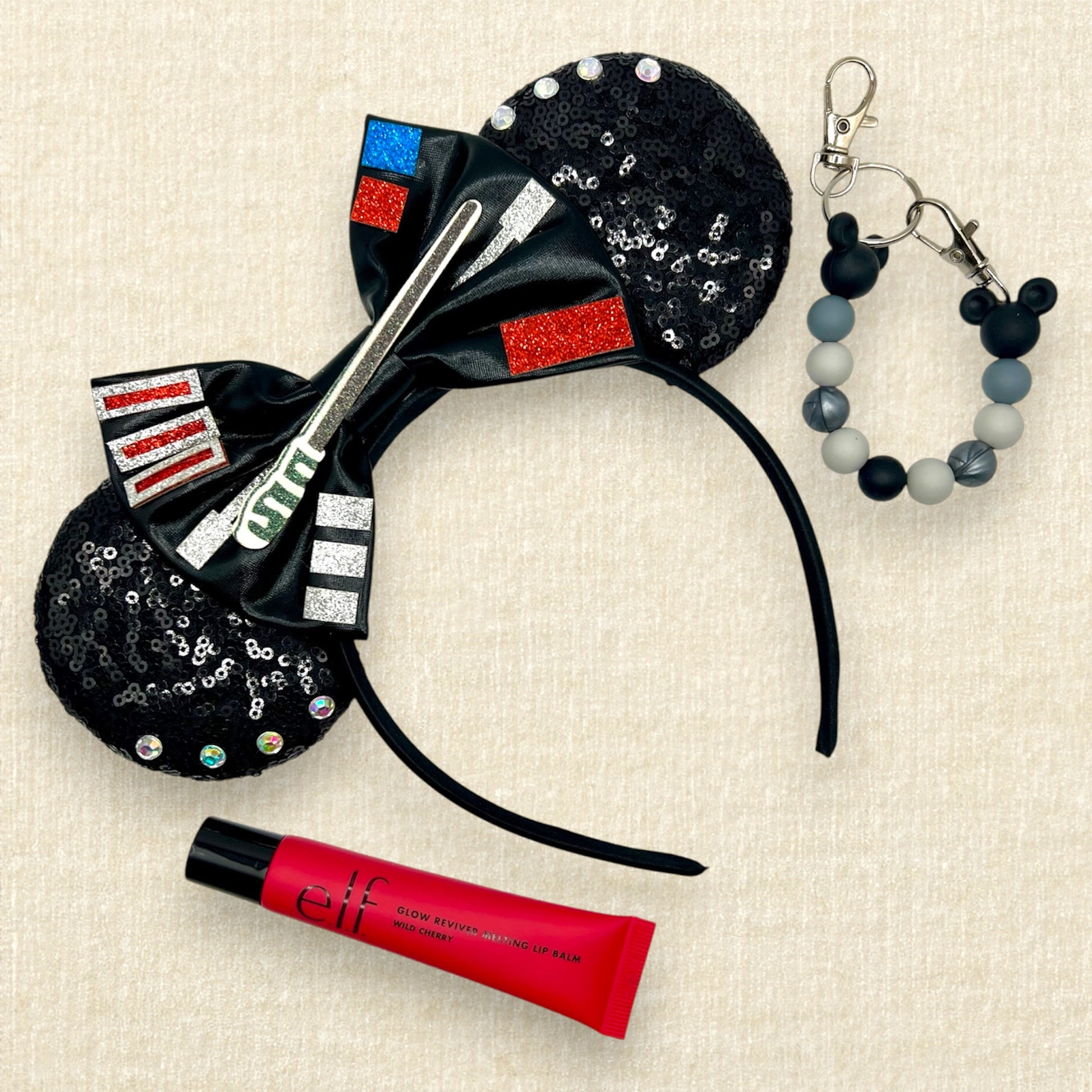 Black sequin mouse ears headband with metallic black bow, glitter details, and rhinestones; red lip gloss; black and silver beaded mouse ears holder clip