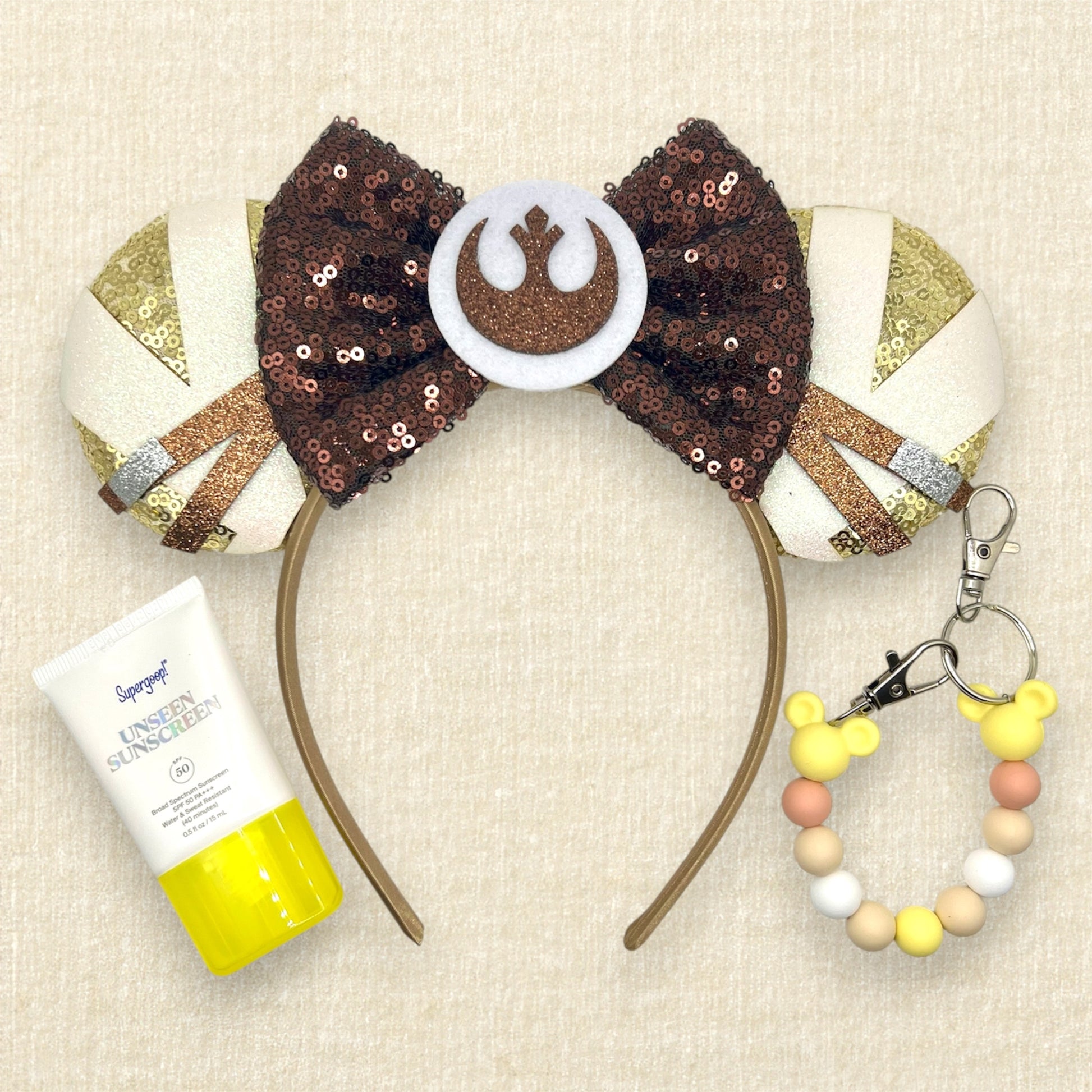 Gold sequin mouse ears with brown and white glitter details, brown sequin bow, and brown glitter emblem; tube of sunscreen; yellow beaded mouse ears holder