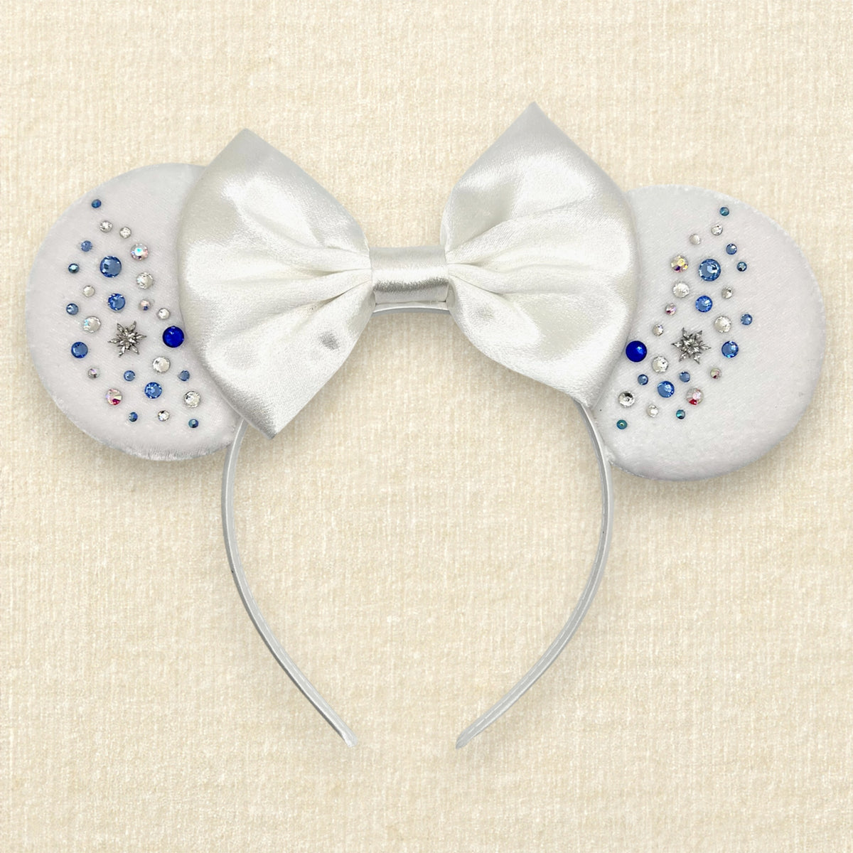 Winter-themed white velvet mouse ears headband with a large white satin bow and decorative blue and silver rhinestones on a textured beige background.