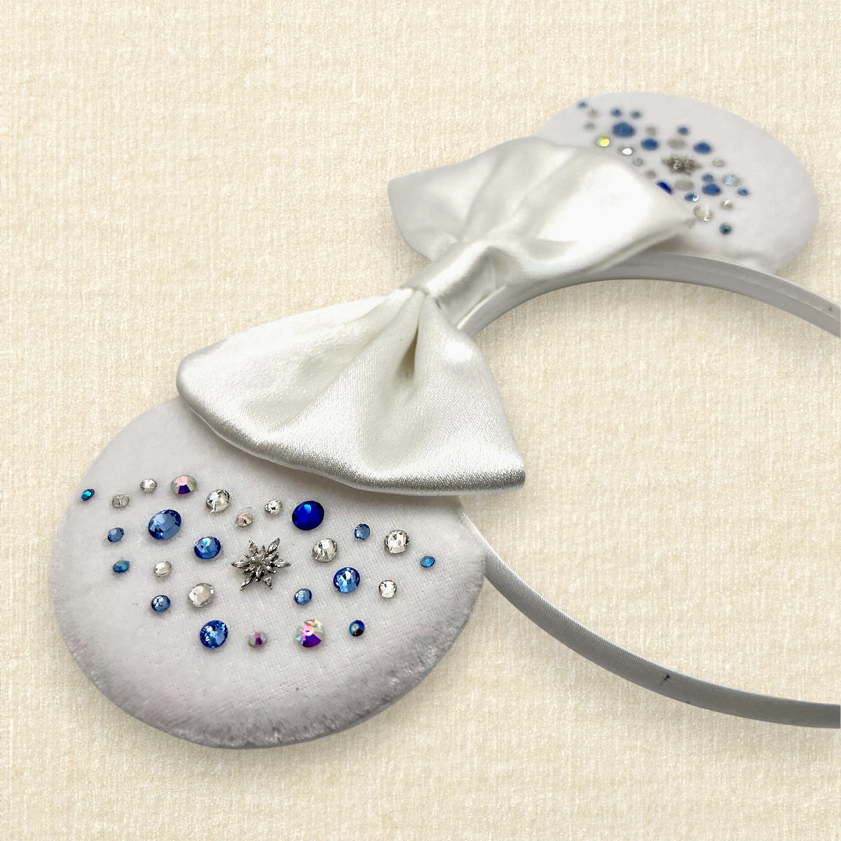 Winter-themed white velvet mouse ears headband with a large white satin bow and decorative blue and silver rhinestones on a textured beige background.