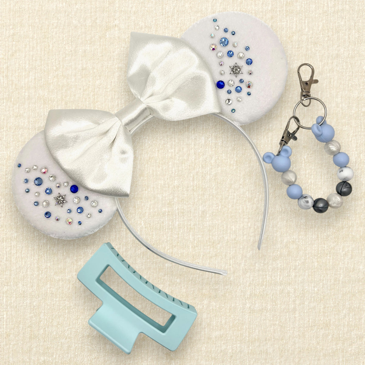 Winter-themed white velvet mouse ears headband with a large white satin bow and decorative blue and silver rhinestones; a light blue claw clip; and a blue, pearl white, and silver beaded mouse ears holder keychain clip