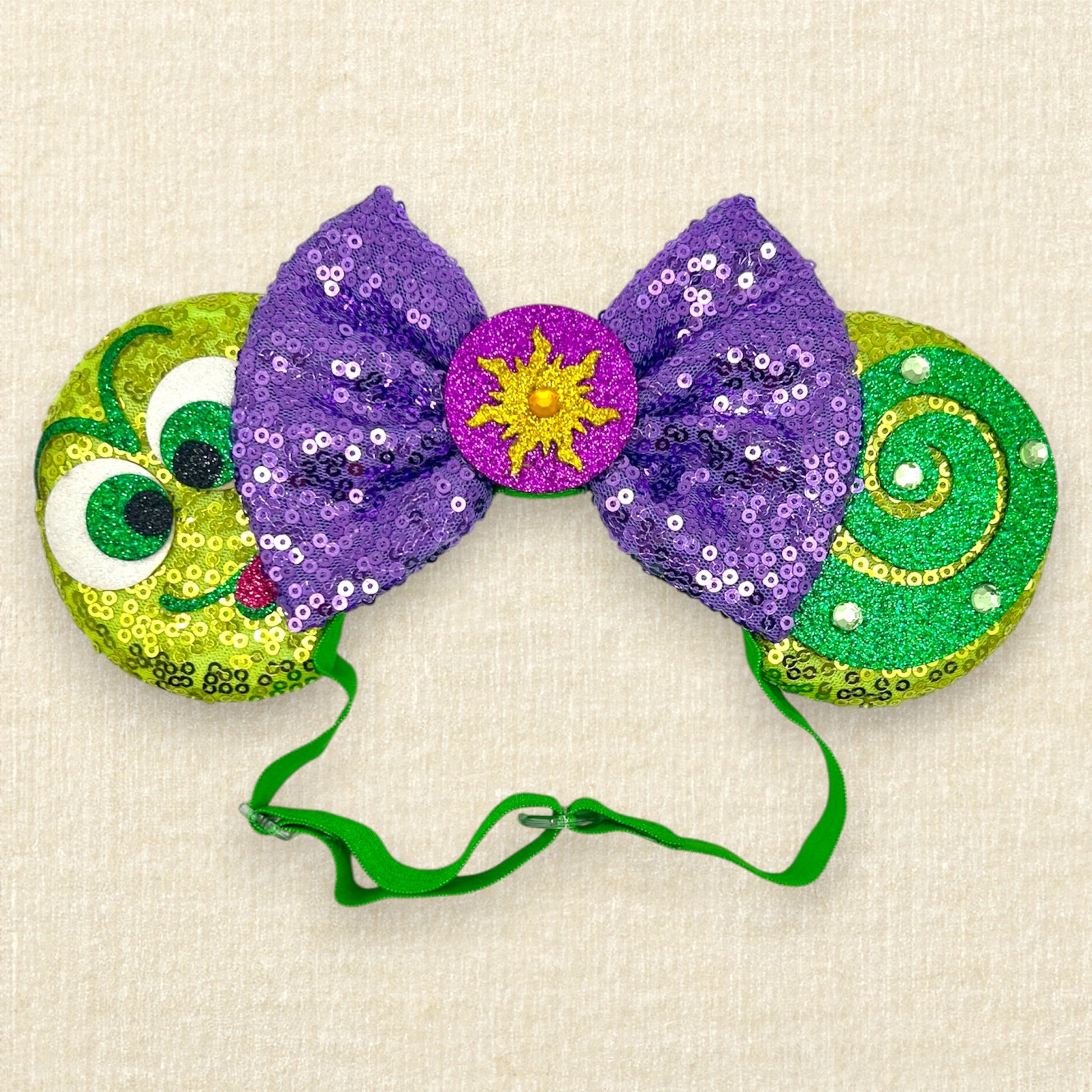Green sequin mouse ears with glitter chameleon details, purple sequin bow with yellow sun centerpiece, and rhinestones on an elastic headband