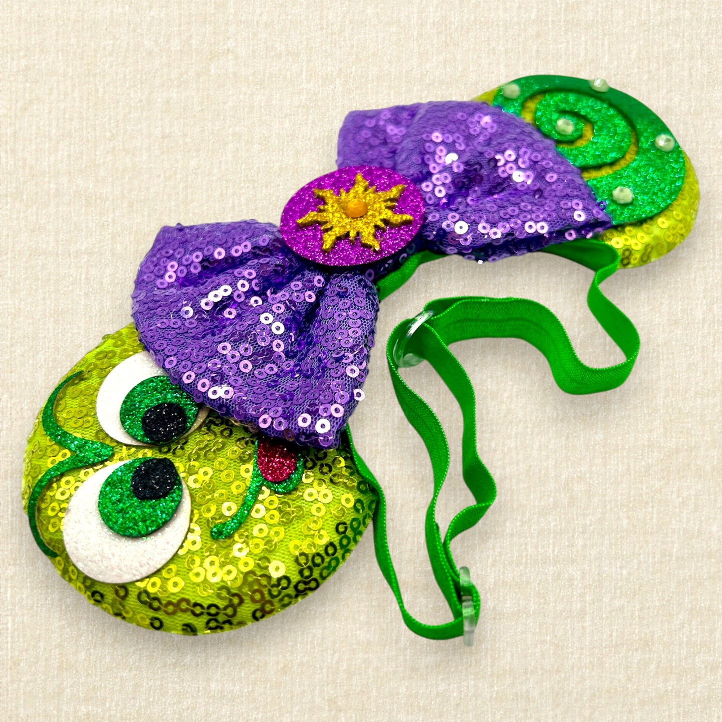 Green sequin mouse ears with glitter chameleon details, purple sequin bow with yellow sun centerpiece, and rhinestones on an elastic headband