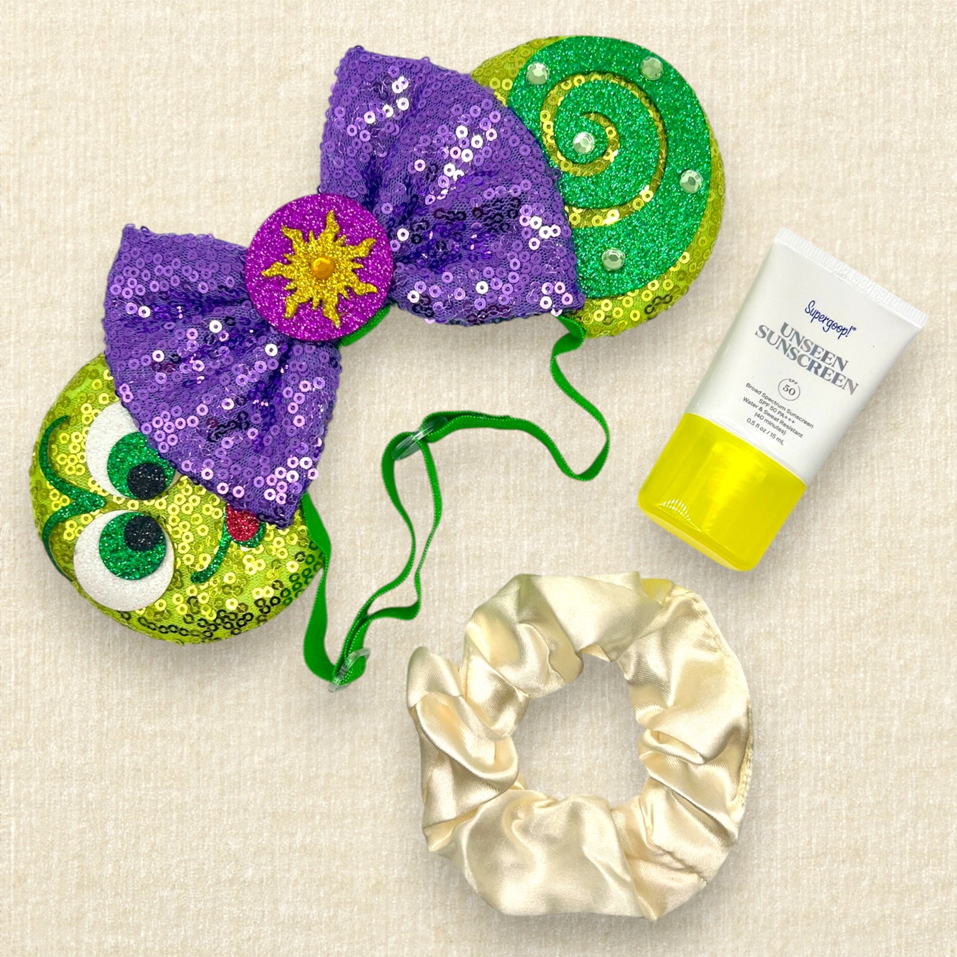 Green sequin mouse ears with glitter chameleon details, purple sequin bow with yellow sun centerpiece, and rhinestones on an elastic headband; tube of sunscreen; cream scrunchie