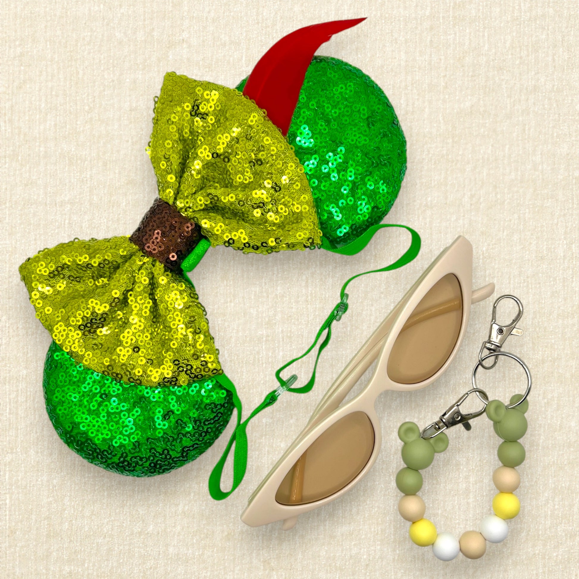 Green sequin mouse ears with lighter green sequin bow and red feather on an elastic headband; cream cat eye sunglasses; green beaded mouse ears holder clip