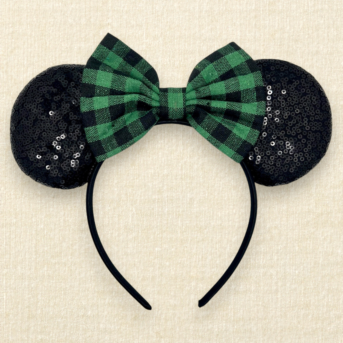 Christmas mouse ears headband with black sequin ears and a green and black plaid bow, displayed on a textured beige background.