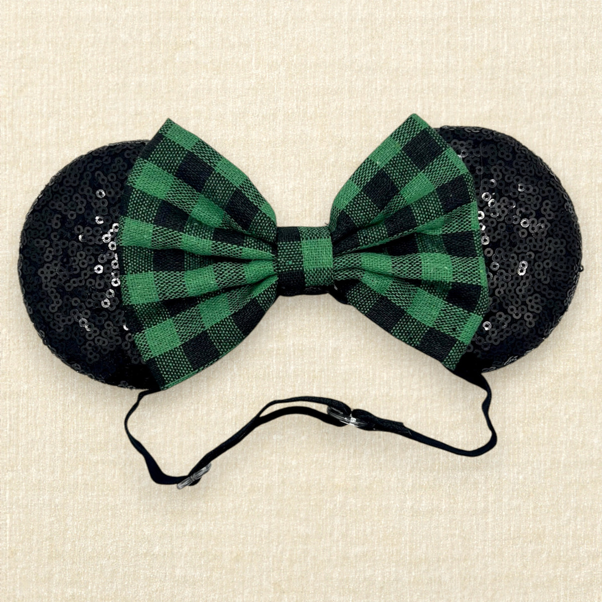 A festive Christmas-themed mouse ears headband with black sequin ears and a green and black buffalo plaid bow on an elastic headband