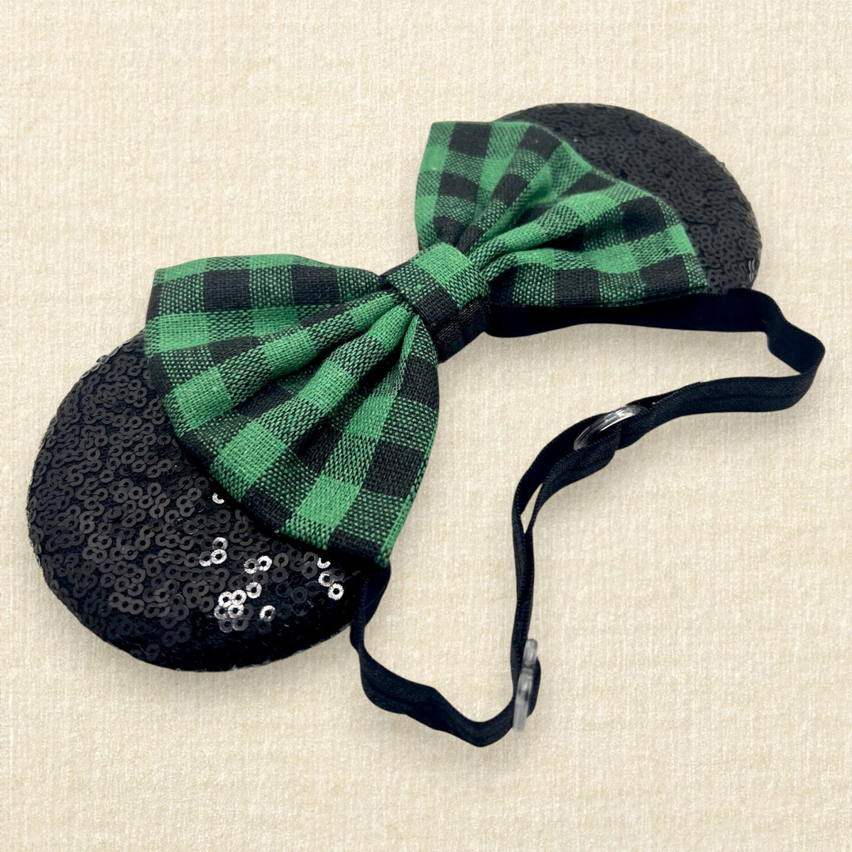 A festive Christmas-themed mouse ears headband with black sequin ears and a green and black buffalo plaid bow on an elastic headband