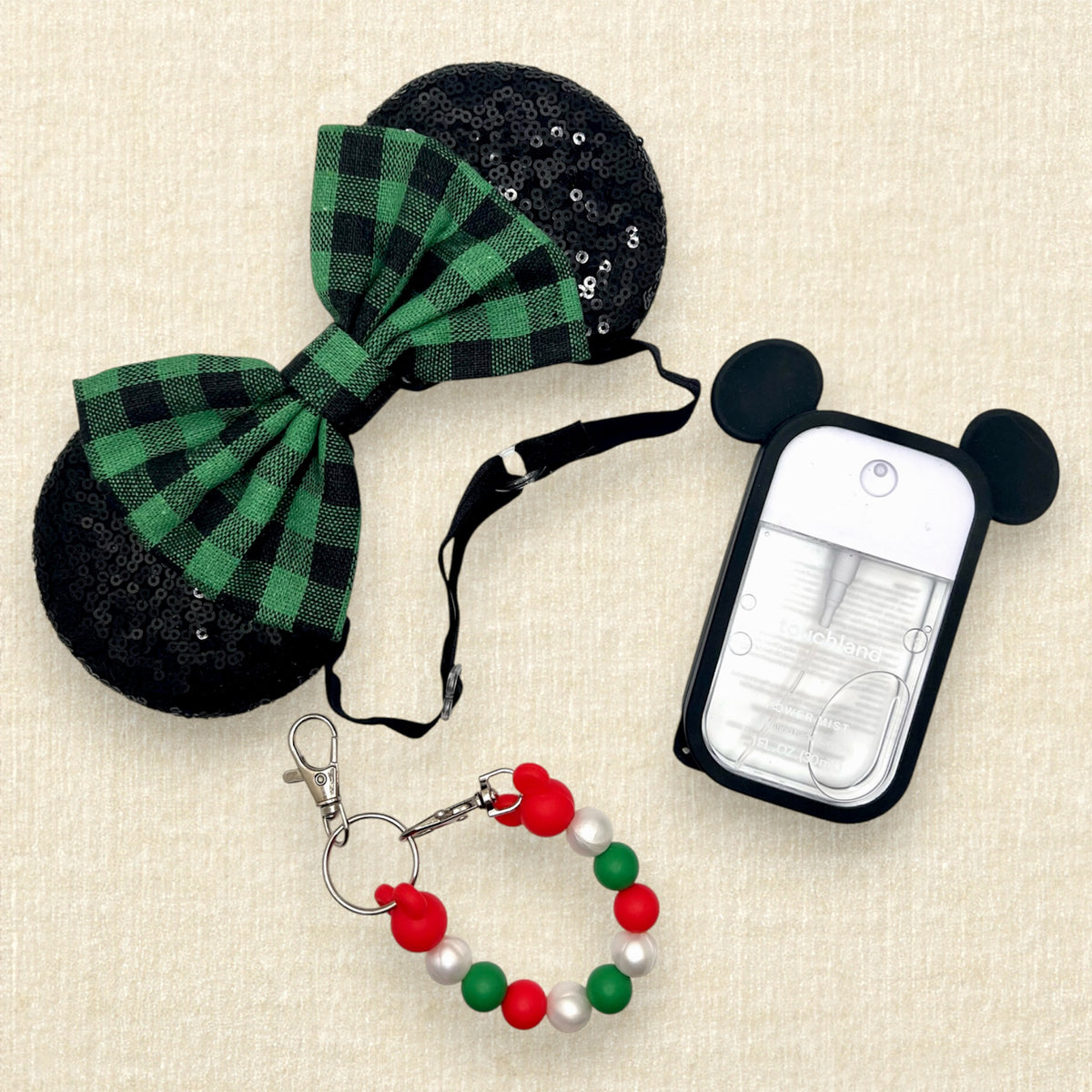 A festive Christmas-themed mouse ears headband with black sequin ears and a green and black buffalo plaid bow on an elastic headband; a red, pearl white, and green beaded mouse ears holder keychain clip; and a spray hand sanitizer with black mouse ears