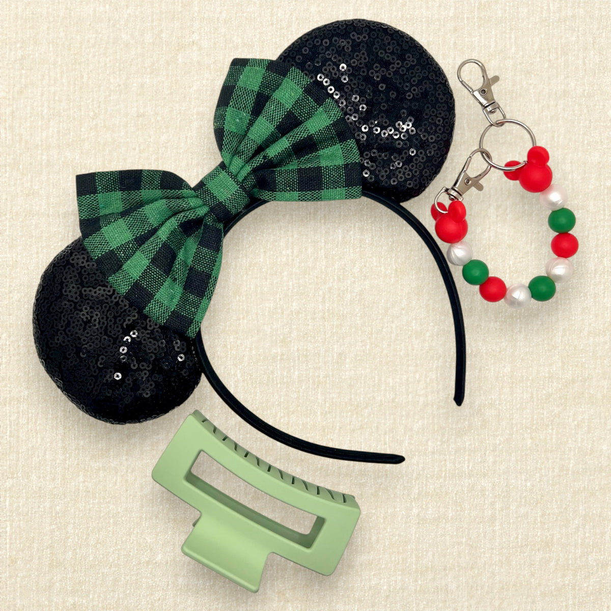 A holiday-themed hair accessory set on a textured cream background, featuring a black sequined headband with a green and black plaid bow, a green hair clip, and a red, green, and white beaded mouse ears holder keychain clip