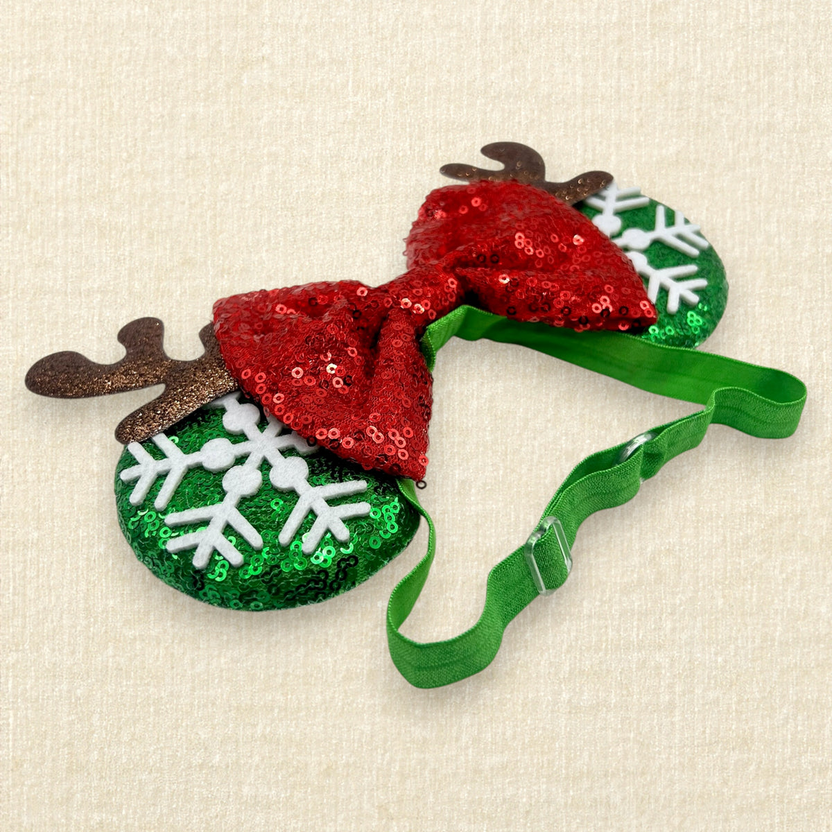 A festive Christmas-themed mouse ears headband with two round green sequin ornaments decorated with white snowflakes and brown reindeer antlers, connected by a red sequin bow and a green elastic strap.