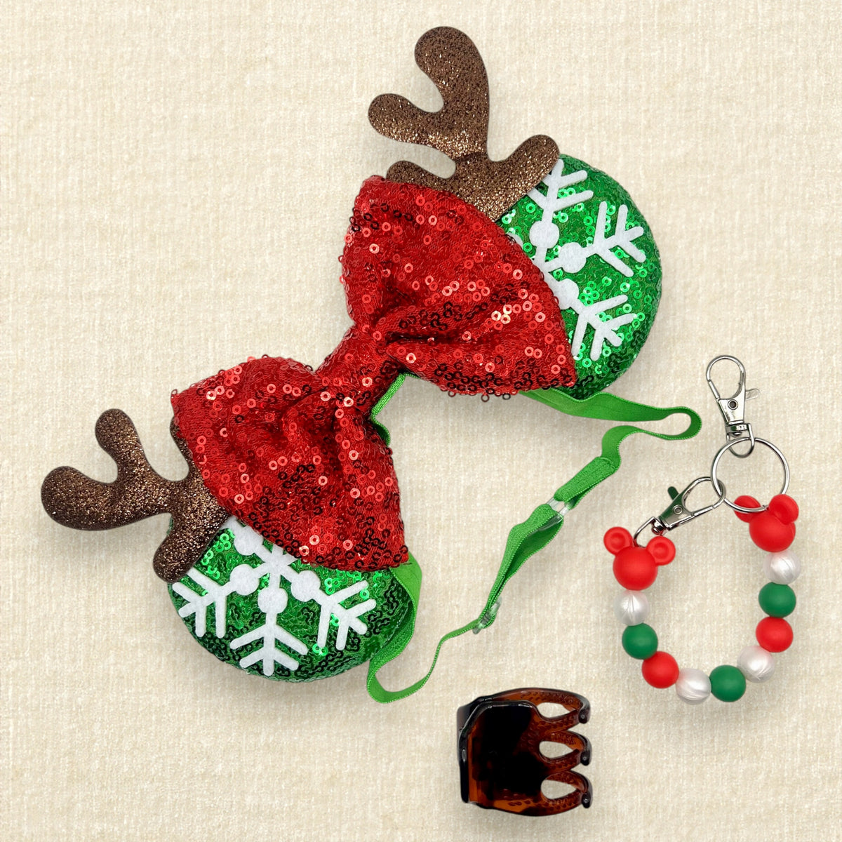 Christmas themed mouse ears headband with green sequin ears, white snowflake details, red sequin bow, and brown glitter reindeer antlers on an elastic headband; red, white, and green beaded mouse ears holder keychain clip; and brown claw clip