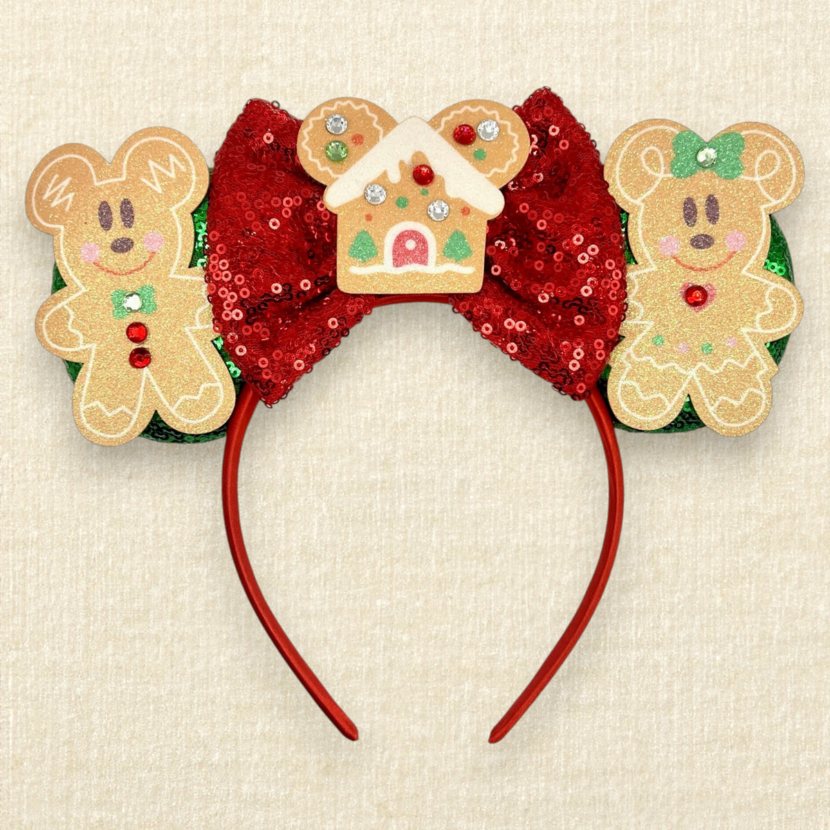 A festive Christmas-themed mouse ears headband featuring gingerbread mouse cookies, a red sequin bow, and a gingerbread house ornament, on a beige textured background.