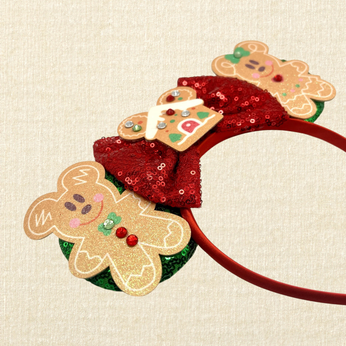 A festive Christmas-themed mouse ears headband featuring gingerbread mouse cookies, a red sequin bow, and a gingerbread house ornament, on a beige textured background.