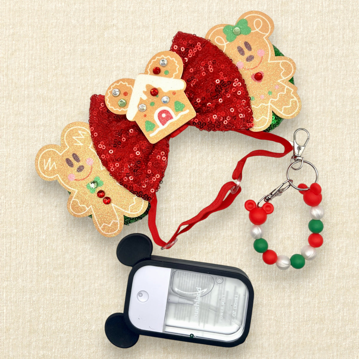 A festive Christmas mouse ears headband featuring two gingerbread mouse cookies, a red sequin bow, and a gingerbread house in the center, with a red elastic headband; a spray hand sanitizer with black mouse ears; and a red, pearl white, and green beaded mouse ears holder keychain clip