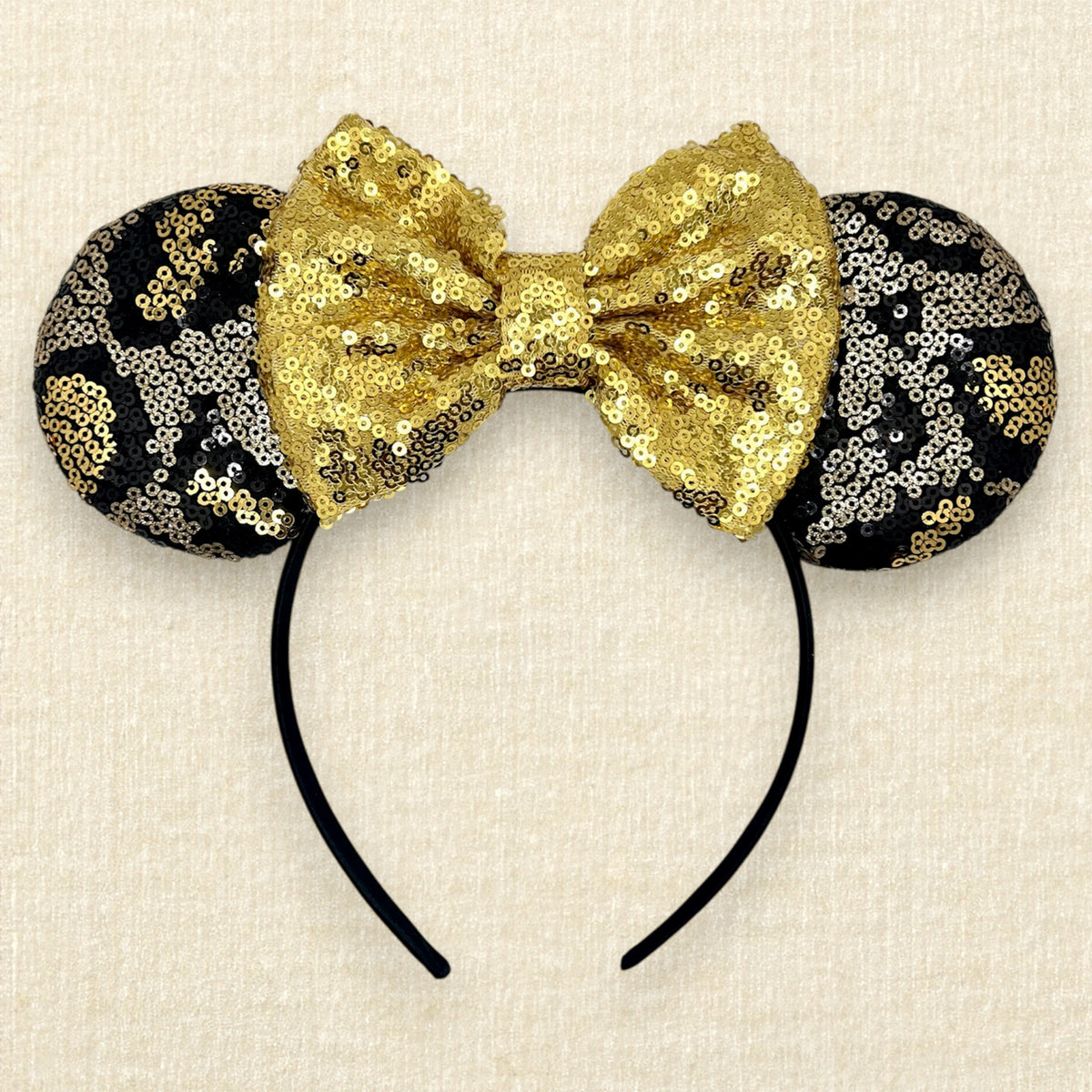 A black and gold animal print sequin mouse ears headband with a large matching gold sequin bow in the center, displayed against a light beige textured background.