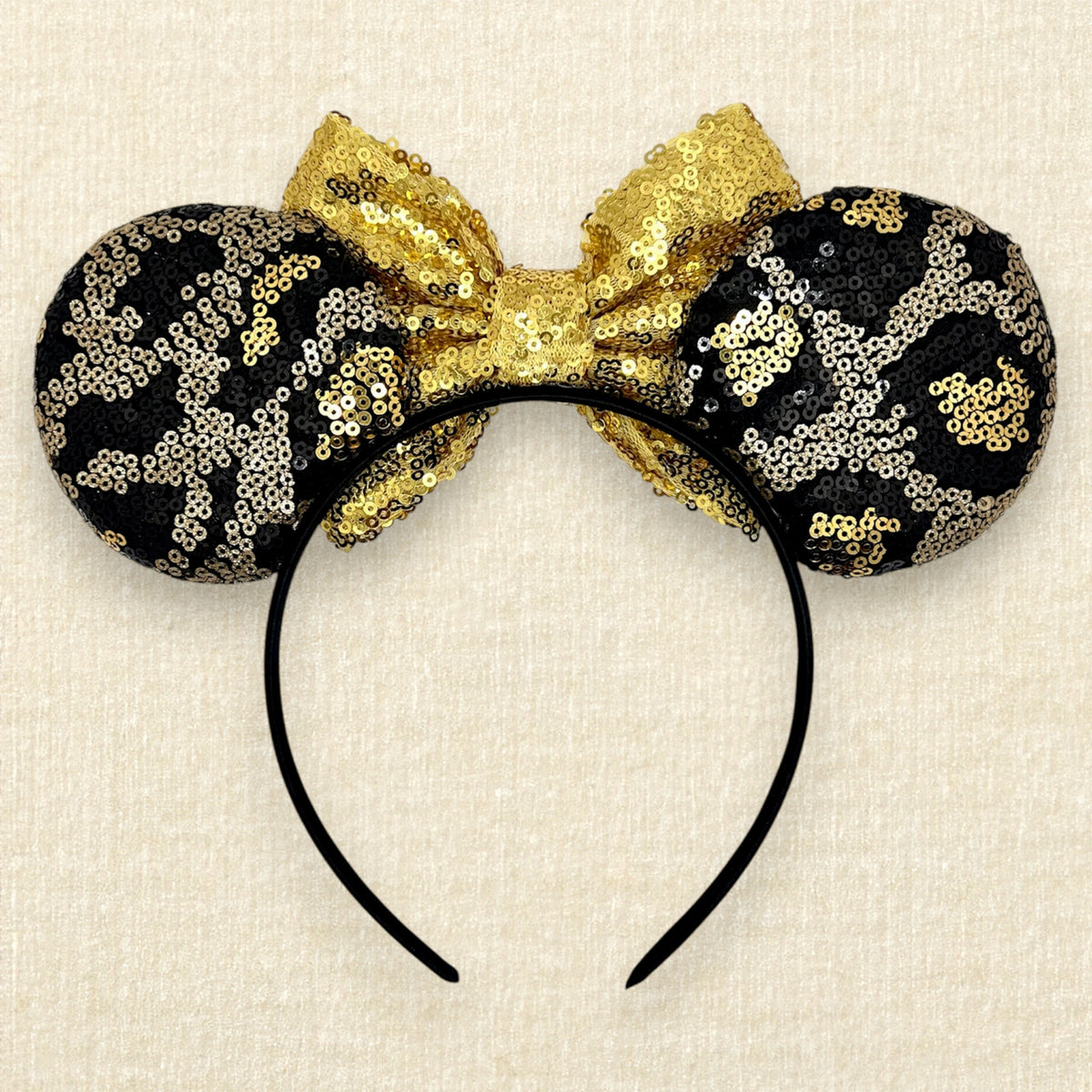 A black and gold animal print sequin mouse ears headband with a large matching gold sequin bow in the center, displayed against a light beige textured background (back view)