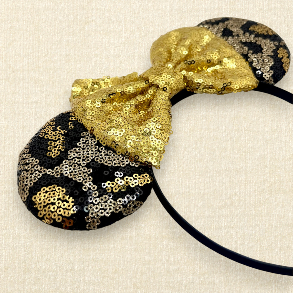A black and gold animal print sequin mouse ears headband with a large matching gold sequin bow in the center, displayed against a light beige textured background.
