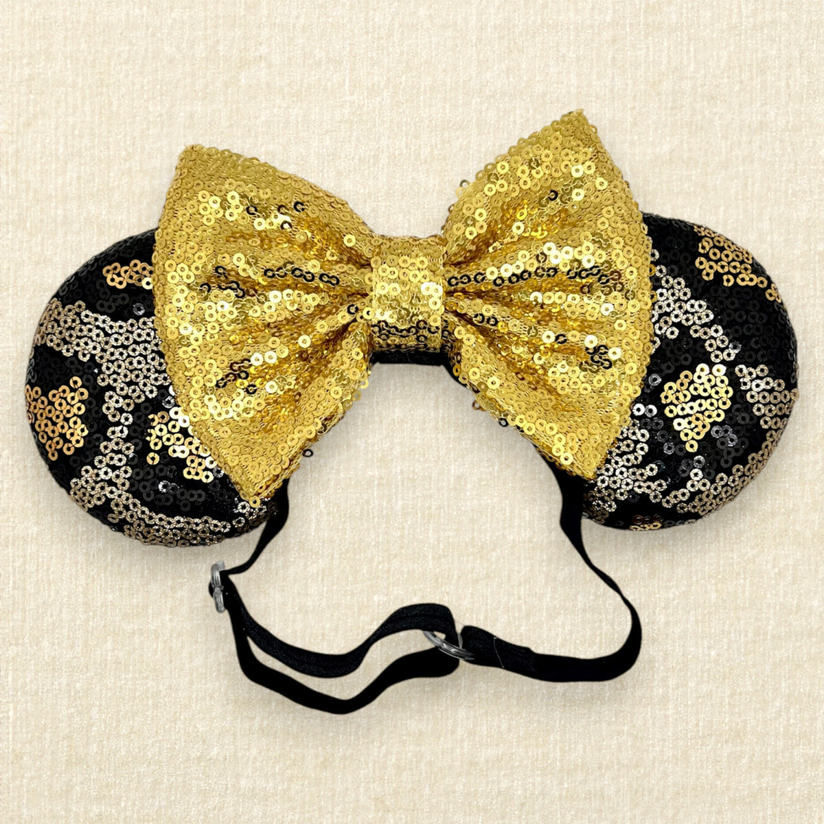 A black and gold animal print sequin mouse ears headband with a large matching gold sequin bow with an elastic headband, displayed against a light beige textured background.