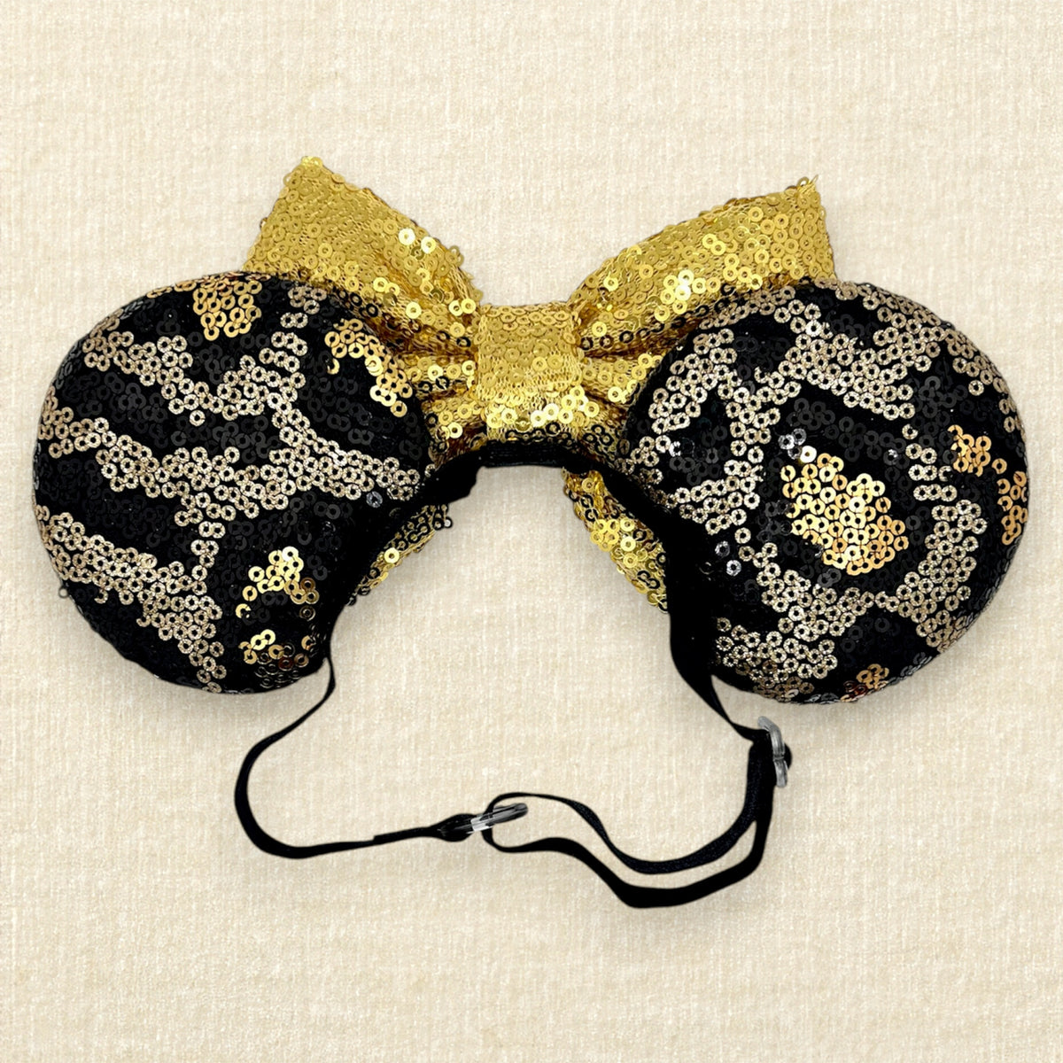 A black and gold animal print sequin mouse ears headband with a large matching gold sequin bow with an elastic headband, displayed against a light beige textured background (back view)