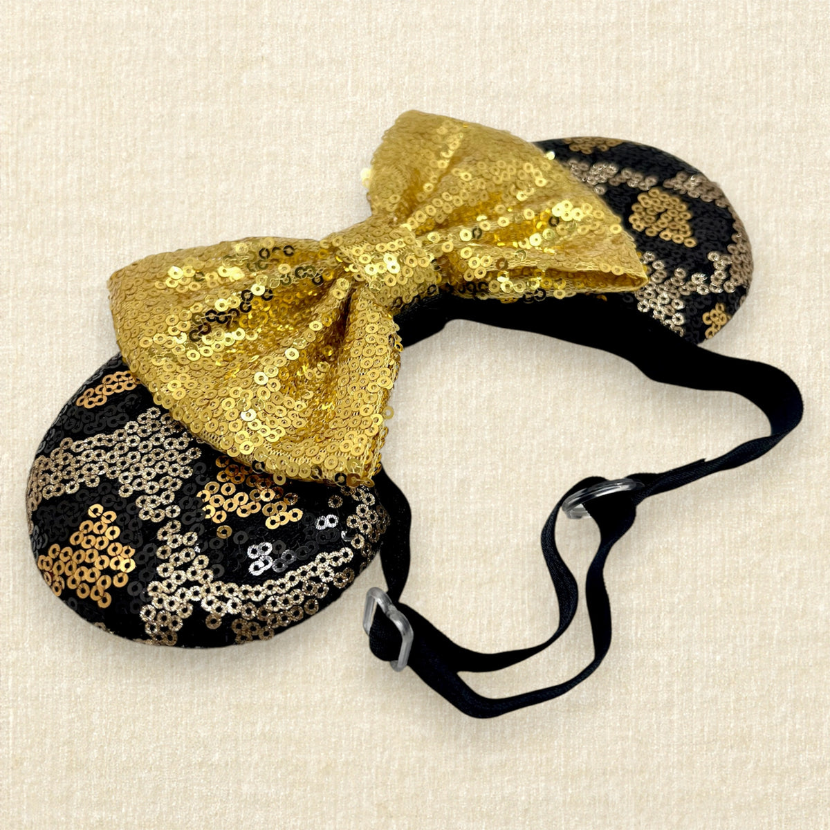 A black and gold animal print sequin mouse ears headband with a large matching gold sequin bow with an elastic headband, displayed against a light beige textured background.