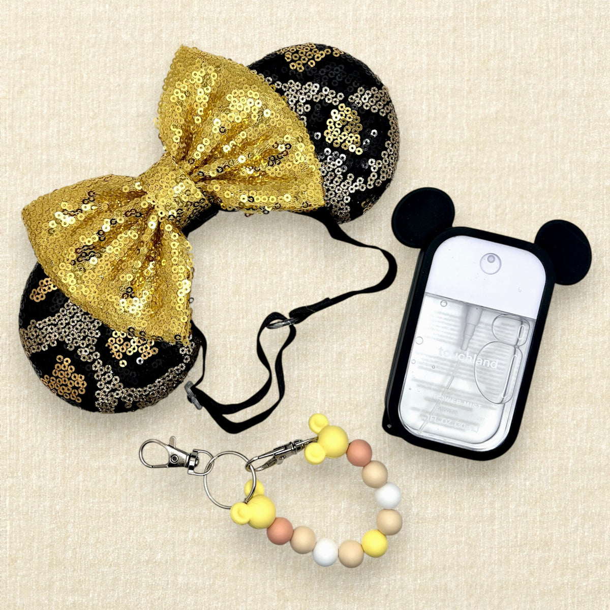 A black and gold animal print sequin mouse ears headband with a large matching gold sequin bow with an elastic headband; a yellow beaded mouse ears holder keychain clip; and a spray hand sanitizer with black mouse ears