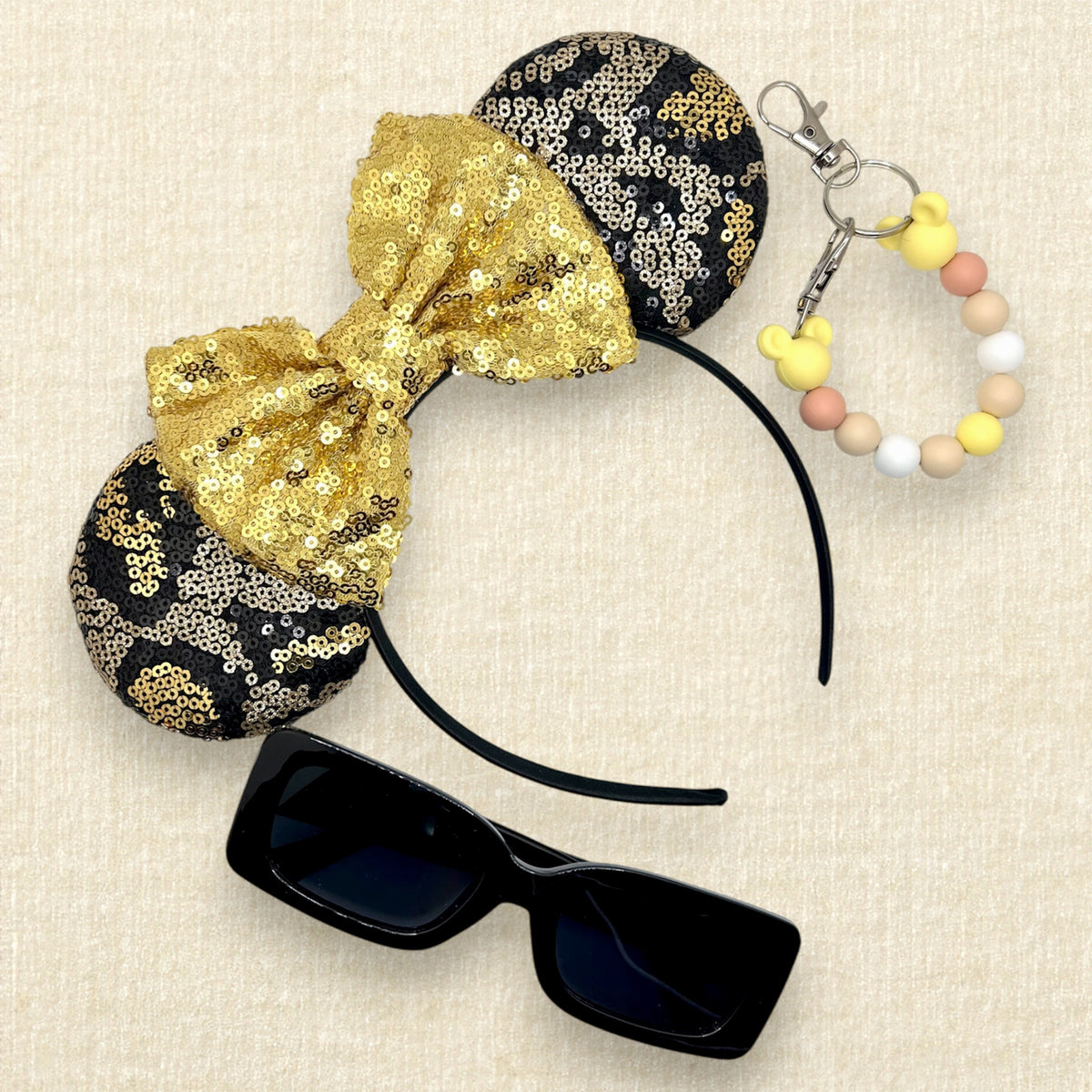 A black and gold animal print sequin mouse ears headband with a large matching gold sequin bow; black sunglasses; and a yellow beaded mouse ears holder keychain clip
