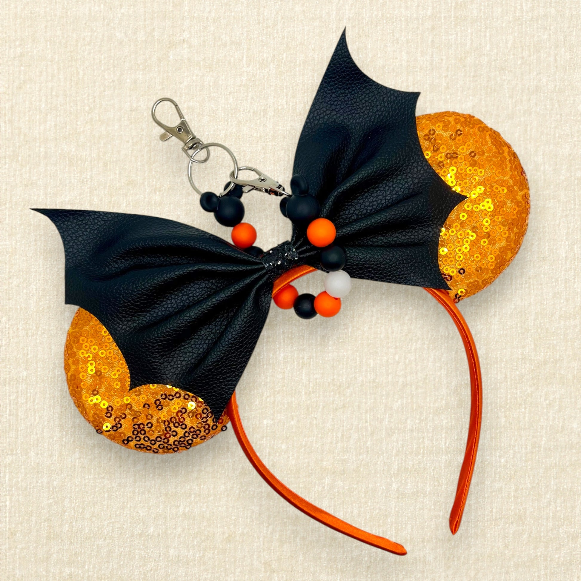 Halloween-themed mouse ears with orange sequin ears and black faux leather bat wing bow, with an orange and black beaded mouse ears holder keychain