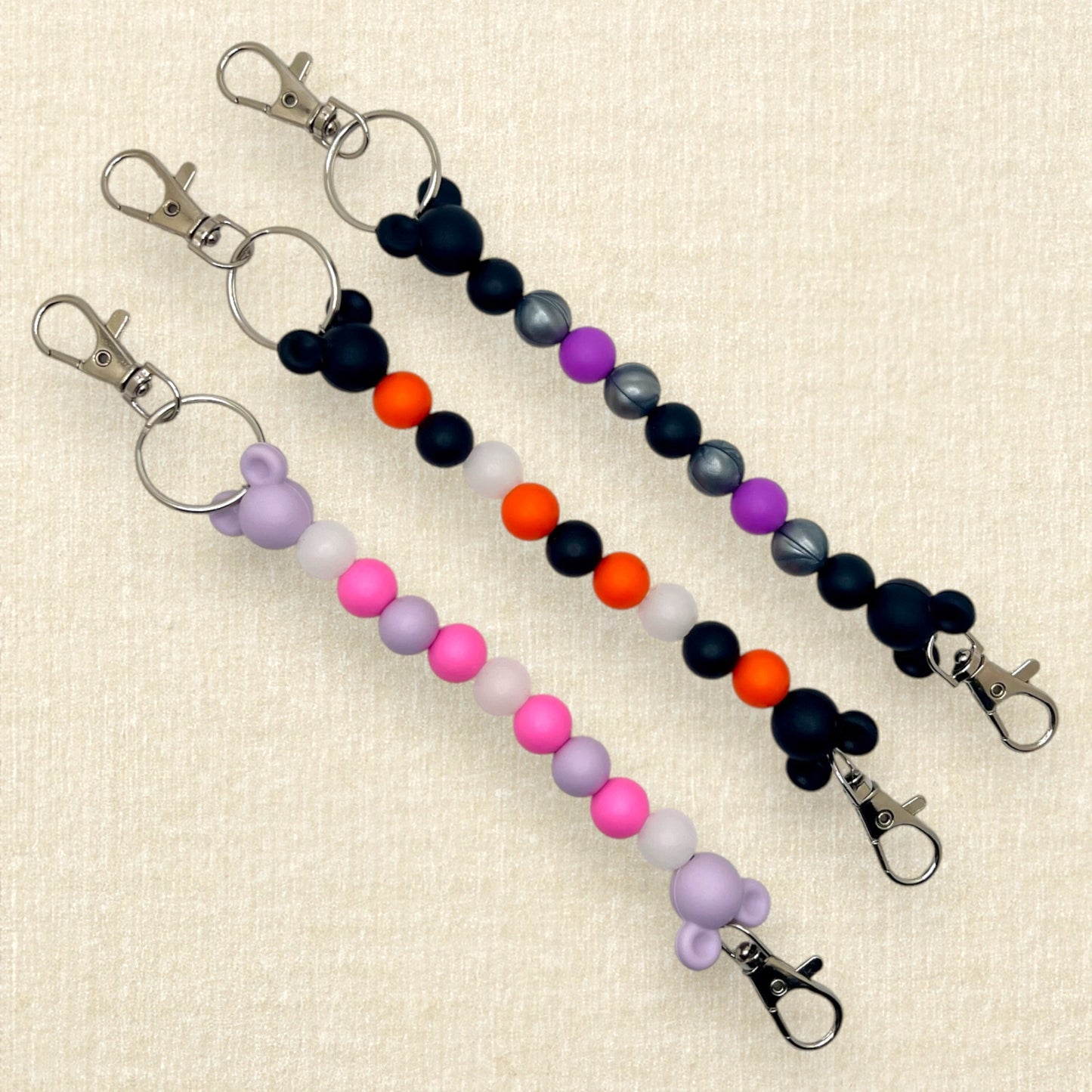 Three beaded mouse ears holder keychains: lavender and pink; orange and black; purple, black, and silver