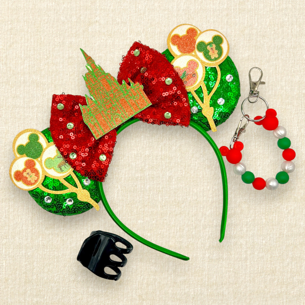 A festive Disney-themed headband with red sequined bow, green sequined ears, red and green glitter castle centerpiece, and Mickey-shaped balloons, shown with a matching red, green, and white beaded mouse ears holder clip and a black hair clip on a cream fabric background.