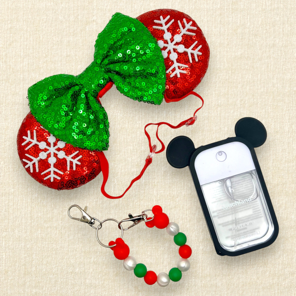A festive Christmas mouse ears headband with red sequin mouse ears, white snowflake details, and a green sequin bow on an elastic headband; red, pearl white, and green beaded mouse ears holder keychain clip; and a white hand sanitizer spray with black mouse ears